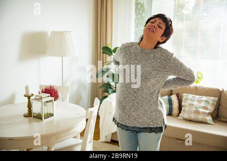 Mature woman suffering from backache at home. Massaging lower back with hand, feeling exhausted, standing in living room. Stock Photo