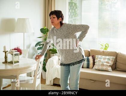 Mature woman suffering from backache at home. Massaging lower back with hand, feeling exhausted, standing in living room. Stock Photo