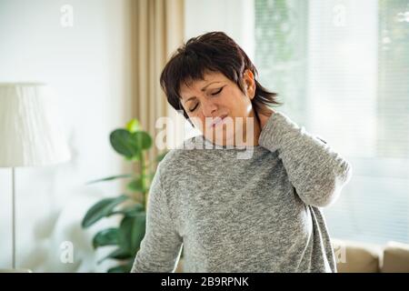 Mature woman suffering from backache at home. Massaging neck with hand, feeling exhausted, standing in living room. Stock Photo