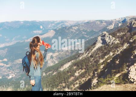 Young beautiful girl travels alone in the mountains in spring or autumn, looks into the distance and enjoys nature, rocks and green forests, view of the landscape. a backpack behind and sportswear, drinking water from an eco bottle Stock Photo