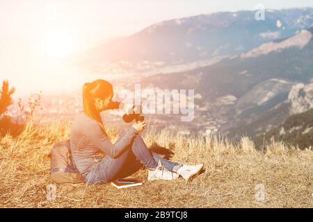 Young beautiful girl travels alone in the mountains in spring or autumn, sits on the edge of the mountain and looks into the distance and enjoys nature, rocks and green forests, view of the landscape. a backpack behind and sportswear, a thermos with a hot drink or tea, freedom and lightness. Back view of woman traveler in cap sitting on mountain top alone and looking at beautiful summer landscape and blue sea view. Drink tea or coffe Stock Photo