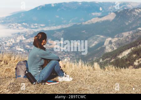 Young beautiful girl travels alone in the mountains in spring or autumn, sits on the edge of the mountain and looks into the distance and enjoys nature, rocks and green forests, view of the landscape. a backpack behind and sportswear, a thermos with a hot drink or tea, freedom and lightness. Back view of woman traveler in cap sitting on mountain top alone and looking at beautiful summer landscape and blue sea view. Stock Photo