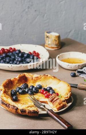 Started eaten fresh baked Dutch baby pancake in ceramic plate with blackberry and red currant berries, bowl of honey, jug of cream, vintage cutlery ov Stock Photo