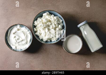 Set of farm dairy produce for breakfast cottage cheese, milk cream, yogurt in ceramic bowls and bottle in row over brown texture background. Flat lay, Stock Photo