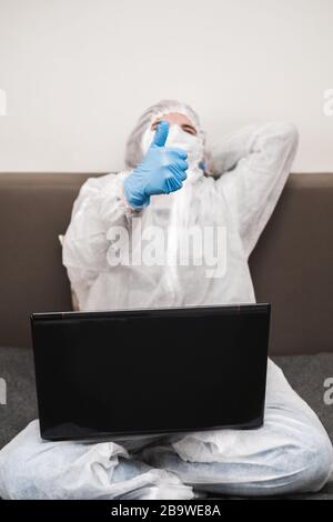 Person in protective white translucent suit, blue rubber gloves, medical mask sits at table at home and works or studies on laptop during quarantine. Stock Photo