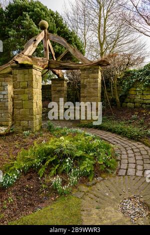 Corner of country cottage garden (rustic arbour, timber beams, stone setts, curving path, millstone, snowdrops on border) - York Gate Garden, Leeds UK Stock Photo