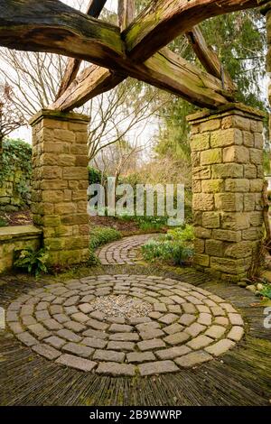 Corner of country cottage garden (rustic arbour with timber beams, stone setts, curving path, circular pattern of stones) - York Gate Garden, Leeds UK Stock Photo