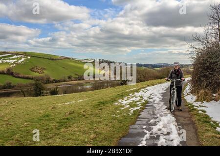 A cyclist on the Dart Valley Cycleway, with unseasonal snow despite sunny weather. Ashsprington near Totnes, Devon, UK. March 2018. Stock Photo