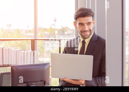 A business man dressed in a neatly dressed suit holds a tablet next to a glass wall in his office with a bright smile and a beautiful orange light. Stock Photo