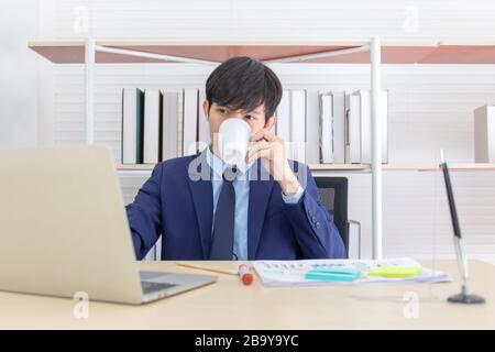 A young Asian businessman sits an afternoon coffee on the desk. Stock Photo