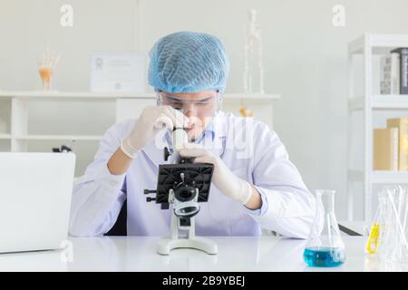 Young Scientist using Microscope in Laboratory. Male Researcher wearing white Coat sitting at Desk and looking at Samples by using Microscope in Lab. Stock Photo