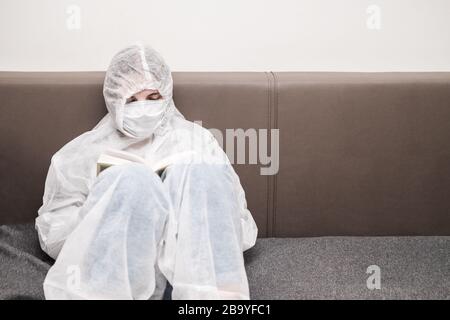 Girl in protective white translucent suit, blue rubber gloves, a medical mask sits on sofa at home and reads book. Stay home during the coronavirus pa