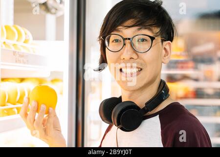 Asian boy with glasses and headphones to listen to music picking up an orange from the fridge of a store Stock Photo