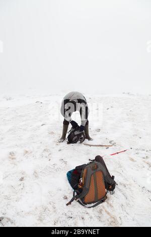 Mountain hiker with a hat and rubber boots at the top of the mountain looking in his backpack and another orange backpack in snow in front of him Stock Photo