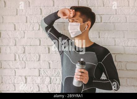 Portrait of tired sports man in medical protective mask on face having rest and drink water after exercising at home. Exhausted fitness man in face ma Stock Photo