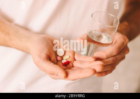 Drug addiction of young man. Sick patient holding oral medicaments and glass of water in hand. Man taking different pills. Treatment of coronavirus CO Stock Photo