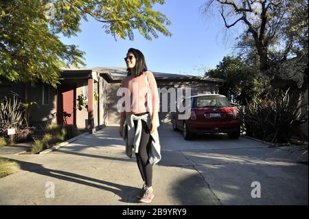 Asian woman wearing a pink sweater walking down a driveway with a tote bag around shoulder. Stock Photo