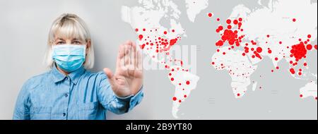 Adult woman in a surgical protective mask shows a stop sign with her hand on the background of foci of infection global map. Protection against virus, SARS, coronavirus epidemic. COVID-19 Stock Photo