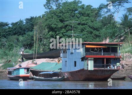 River craft of all sizes moored along the Mekong River in Laos, near Huay Xai. The crew of the largest - a 'slow boat' - will live in the accommodation on the stern. On the bank, men are building a new boat. Stock Photo