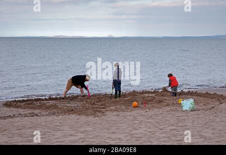 Portobello, Edinburgh, Scotland, UK. 25th March 2020. Extremely quiet afternoon  on Portobello  Beach, pictured this father and sons occupy themselves by building a large sand structure. Temperature of 13 degrees centigrade. Stock Photo