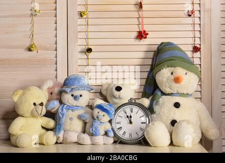 Toys placed on chest of drawers on wooden wall and garlands background. Decorations in festive room. Celebration and New Year decor concept. Snowmen and teddy bears near oldfashioned alarm clock Stock Photo