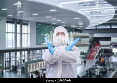 man epidemiologist in coverall disposable anti-epidemic antibacterial isolation suit shows a prohibition gesture against the interior of the airport o Stock Photo