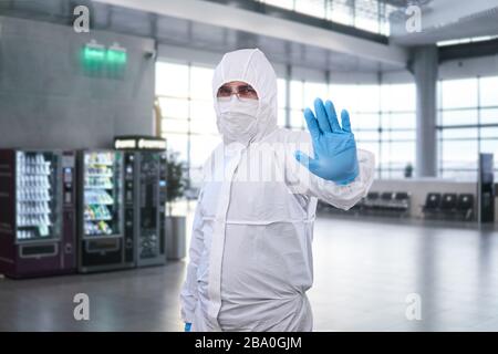 male epidemiologist in coverall disposable anti-epidemic antibacterial isolation suit shows a prohibition gesture against the waiting room at the airp Stock Photo