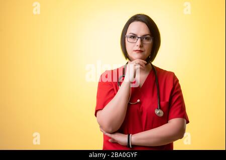 Portrait of beautiful woman doctor with stethoscope wearing red scrubs, holding hand under her chin, having doubtful posing on a yellow isolated backg Stock Photo