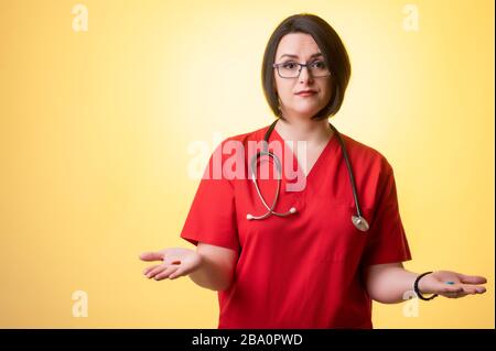 Portrait of beautiful woman doctor with stethoscope wearing red scrubs, showing red and blue pills on hand in doubt, posing on a yellow isolated backg Stock Photo