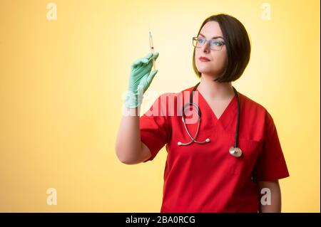 Portrait of beautiful woman doctor with stethoscope wearing red scrubs, with medical glove and syringe in hand posing on a yellow isolated background. Stock Photo