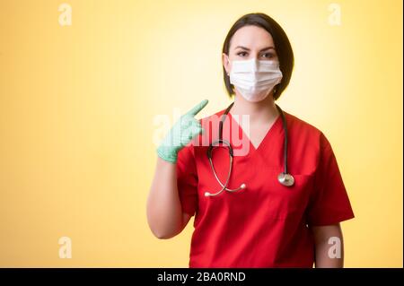 Portrait of beautiful woman doctor with stethoscope wearing red scrubs, wears a protective mask, with her fingers pointed posing on a yellow isolated Stock Photo