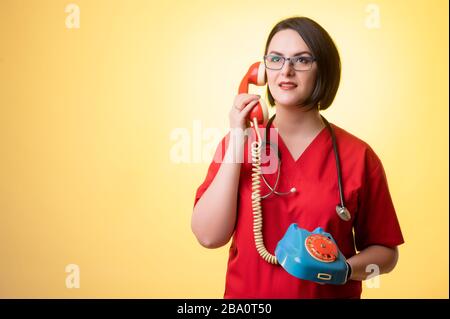 Portrait of beautiful woman doctor with stethoscope wearing red scrubs, with brown hair, displeased face and gestures and talking on old phone, posing Stock Photo