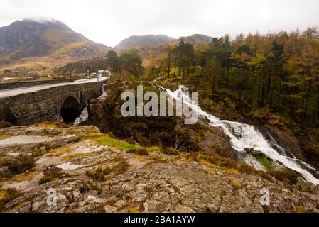 Rugged Welsh landscape, with a road and bridge over fast flowing river, with a waterfall and autumn colors. There is a pine forest and low cloud over Stock Photo