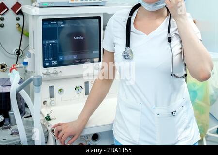 An anesthetist in white medical clothes, a mask and a stethoscope on his neck is standing in the operating room. Behind the doctor are monitors of hig Stock Photo
