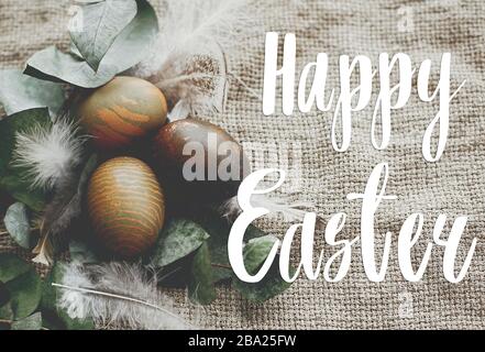Happy Easter text. Easter greetings card. Stylish easter eggs with feathers on rustic table. Natural dyed green easter eggs with eucalyptus branch, sp Stock Photo
