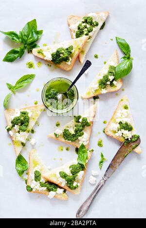 Crispy toasts (crostini, bruschetta) with cottage cheese (ricotta) and freshly prepared pesto. Homemade basil pechto sauce in a jar on a light backgro Stock Photo