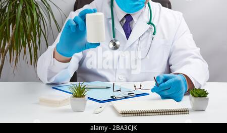 male doctor in a white coat and blue latex gloves sits at a white desk in his office and holds a plastic white jar of pills, concept for receiving pat Stock Photo