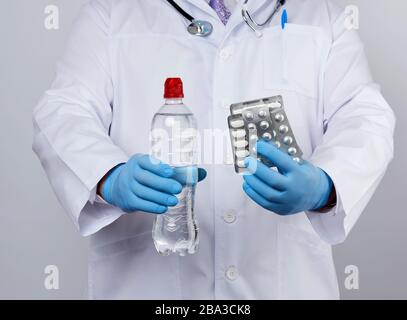 adult doctor therapist is dressed in a white uniform coat and blue sterile gloves is standing and holding a stack of pills in blister packs, white bac Stock Photo