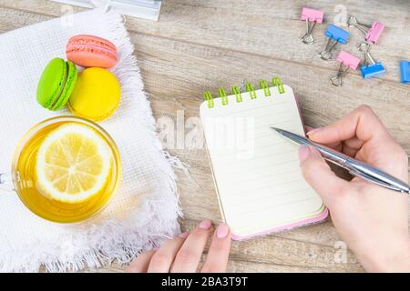 The hand holds a silver pen and writes in an open notebook, next to a notebook on a white towel there is mint tea with lemon and multi-colored muffins Stock Photo