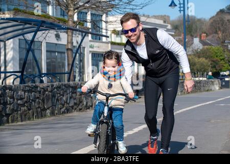 Young girl being taught to ride a bike by her father during a daily exercise excursion on the usually-busy seaside promenade at Mumbles. A Government Stock Photo