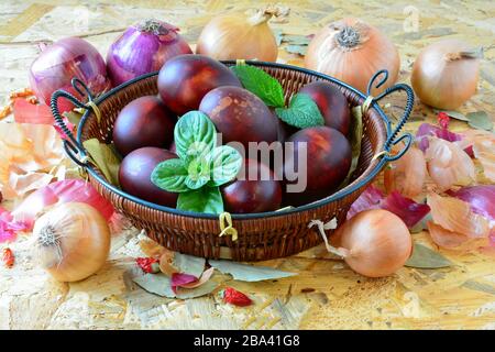 Easter eggs in wicker basket and yellow and red onion, peppers and green basil and lemon balm leaves on wooden background, close up Stock Photo