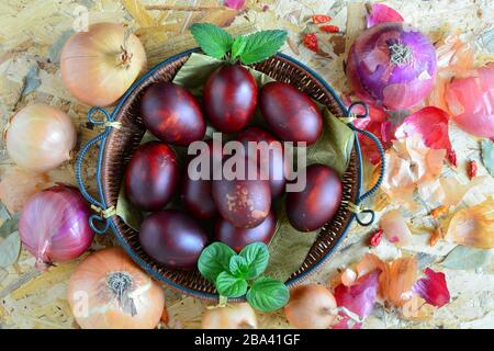 Easter eggs in wicker basket and yellow and red onion, peppers and green basil and lemon balm leaves on wooden background, top view Stock Photo
