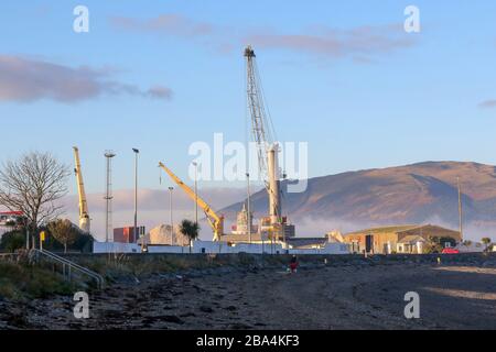 Person with dogs walking along the beach at Greenore with the industrial port and cranes of Greenore Port working in the background on a winter's day. Stock Photo