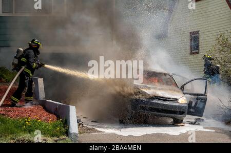 Two firemen fighting a fire in a car. One is spraying the car with a hose, both are wearing oxygen tanks and masks.  Fire visible in the window. Stock Photo