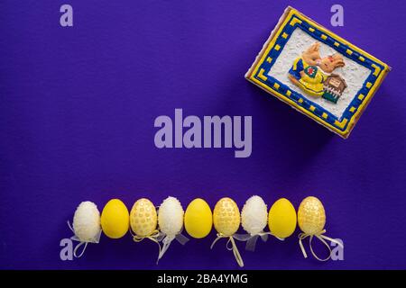 White and yellow fake plastic easter eggs and a closed bunny box on a dark purple surface Stock Photo