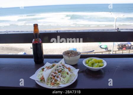 MEXICAN FOOD PLATE WITH BAJA STYLE FISH TACOS WITH A BEER, TOMATO SAUCE AND LEMONS IN THE BACKGROUND SERVED IN FRONT OF THE SEA Stock Photo