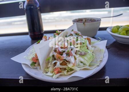 MEXICAN FOOD PLATE WITH BAJA STYLE FISH TACOS WITH A BEER, TOMATO SAUCE AND LEMONS IN THE BACKGROUND SERVED IN FRONT OF THE SEA Stock Photo