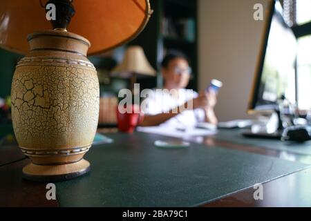 Work from home concept. Focus on table lamp with blurred home background and man on cell phone. Stock Photo
