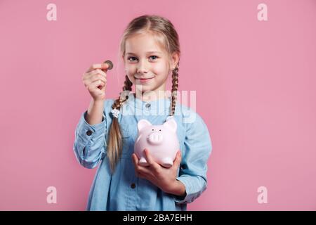 Little girl saving money in a piggybank on pink background Stock Photo