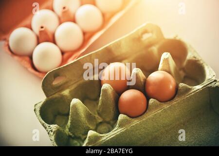 Raw eggs in white and brown shells lie in red and green cardboard packages, standing on a white table and illuminated by sunlight. Stock Photo